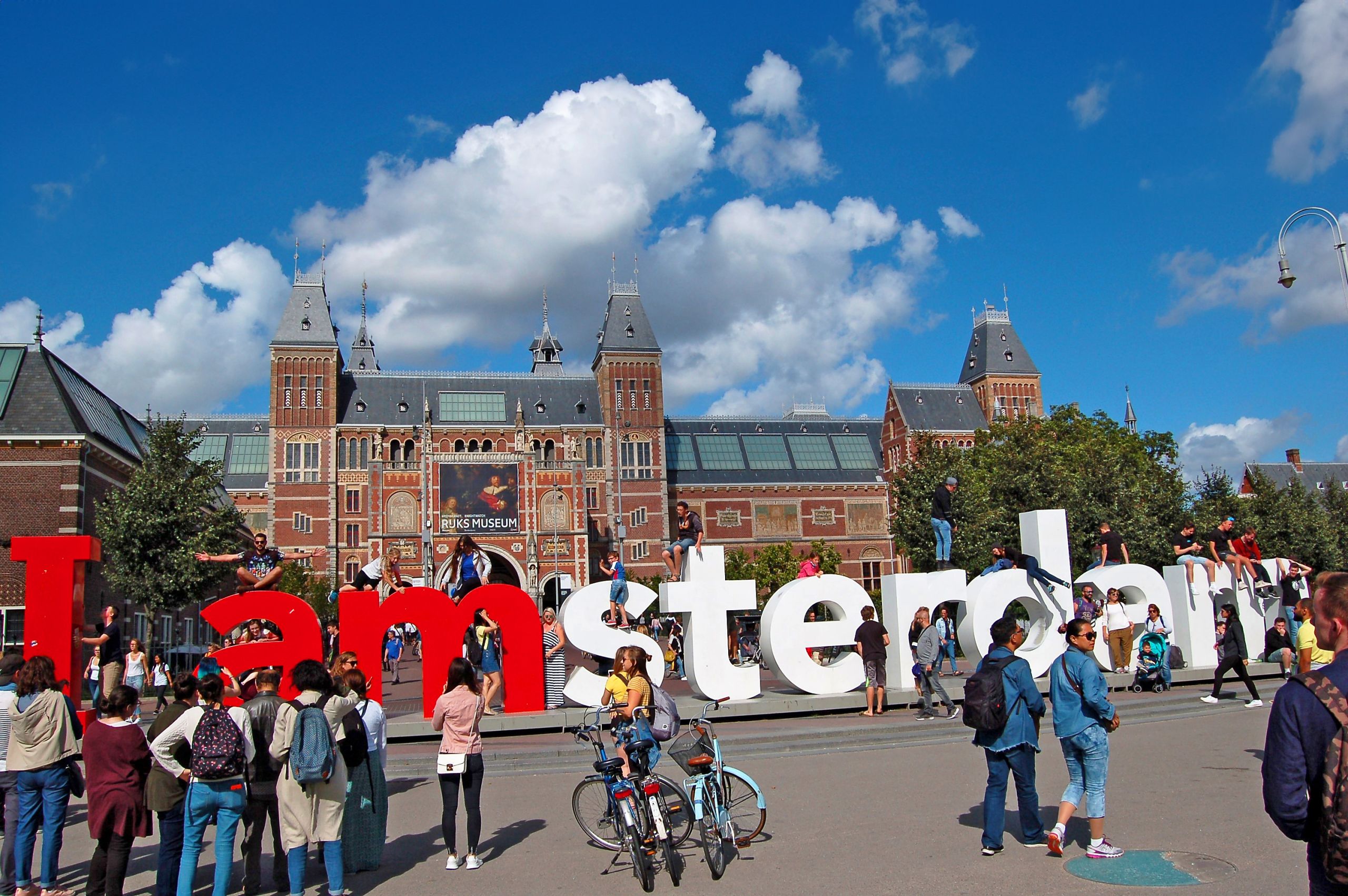 Cyclists posing at the red “I amsterdam” sign in front of the Rijksmuseum, Museumplein – Amsterdam, Netherlands